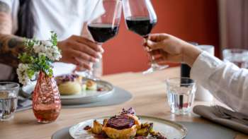 Zwei Personen stoßen mit Rotwein an, im Vordergrund ein liebevoll angerichtetes Hauptgericht