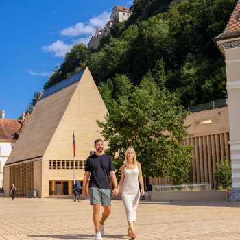 Ein Paar spaziert über den sonnigen Regierungsplatz in Vaduz mit Blick auf das moderne Landtagsgebäude und das Schloss Vaduz im Hintergrund.