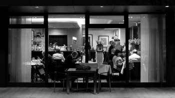 Schwarz-weiß Foto eines belebten Restaurants mit Blick von außen durch die Glasfront.