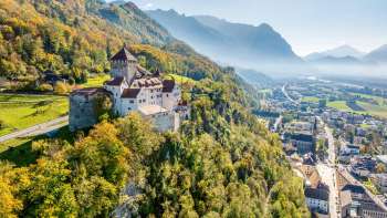 Luftaufnahme von Schloss Vaduz auf einem Hügel über der Stadt, mit Alpenpanorama und Blick ins Rheintal bei klarer Sicht.