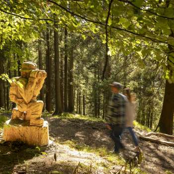 Wanderer passieren eine geschnitzte Figur mit Fernglas am WalserSagenWeg