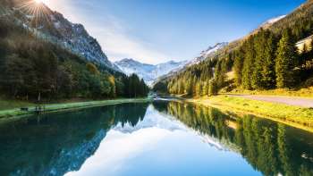 Spiegelglatter Gänglesee in Steg, umrahmt von dichten Wäldern und schneebedeckten Bergen bei Sonnenschein