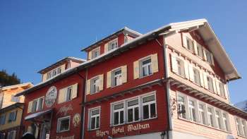 Außenansicht des Alpenhotel Vögeli Malbun mit der Elchbar im Erdgeschoss – rotes Traditionshaus mit weißer Beschriftung vor blauem Himmel.