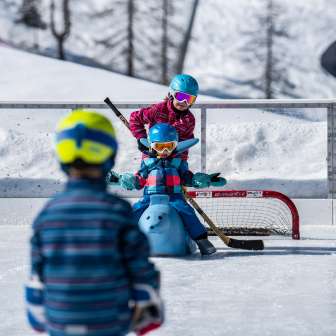 Kinder spielen mit Helm und Hockeyschläger auf dem Eisplatz in Malbun, ein Kind sitzt auf einem Eislauf-Hilfstier vor einem Tor