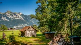 Kleine Holzscheune auf einer Wiese mit Feuerholzstapel und Blick auf die Alpen entlang der Route 66 in Liechtenstein