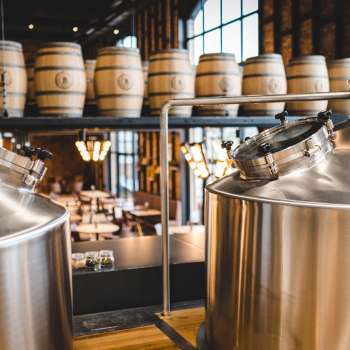 Blick in eine moderne Brauerei mit glänzenden Edelstahltanks und gestapelten Holzfässern im Hintergrund.