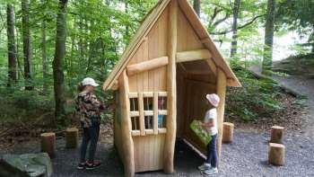 Zwei Kinder erkunden eine kleine Holzhütte im Wald, die Teil des Erlebniswegs „Wurz und Zirp auf Zeitreise“ in Liechtenstein ist – ein Naturabenteuer zum Anfassen.