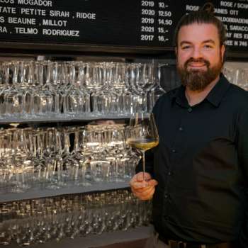 Sympathischer Barkeeper der WY Weinbar mit Weinglas vor stilvollem Regal voller Gläser und Weine.