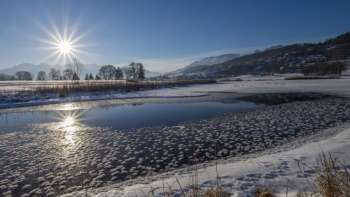 Sonnenaufgang am Egelsee in Mauren im Winter mit glitzernder Eisfläche, Schnee und sonniger Bergkulisse.
