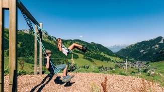 Zwei Kinder schaukeln auf einem Bergspielplatz mit Blick über das Sommerpanorama von Malbun in Liechtenstein