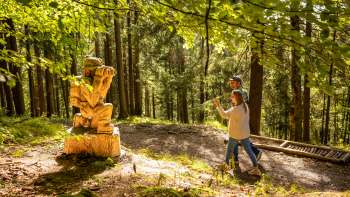 Frau zeigt auf eine detailreiche Holzskulptur im Wald am WalserSagenWeg
