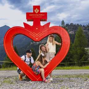 Familie posiert am roten Herzrahmen entlang des Alpherzwegs in Malbun, Liechtenstein, umgeben von alpiner Natur und Bergen.