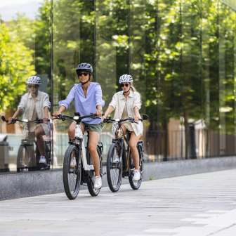 Zwei Frauen auf E-Bikes fahren durch das moderne Zentrum von Vaduz in Liechtenstein, mit Glasfassade und urbanem Flair
