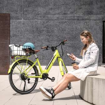 Junge Frau sitzt mit Kaffee und Smartphone neben einem gelben E-Bike im urbanen Zentrum von Vaduz