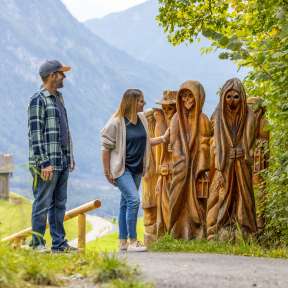 Paar betrachtet geschnitzte Holzfiguren am Walser Sagenweg in Triesenberg mit Blick ins Tal und Bergen im Hintergrund