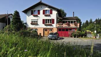 Charmante Außenansicht des Bed & Breakfast Büchel mit roten Fensterläden und Blumenwiese im Vordergrund.