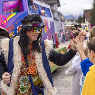 Hippie verkleidet bei Kinderfasnacht in Mauren, High-Fives mit Kindern vor buntem Umzugswagen.