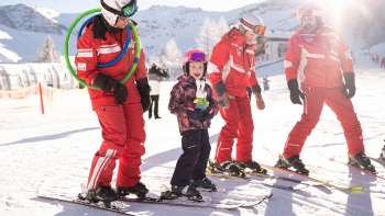 Drei Skilehrer in roten Anzügen und ein Kind auf Skiern im Schnee, im Hintergrund verschneite Berge und Sonnenschein