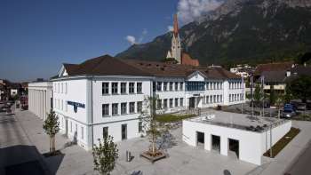 Das Domus-Gebäude in Schaan mit weißer Fassade und Blick auf die Pfarrkirche – ein kulturelles Zentrum und Begegnungsort im Herzen von Liechtenstein