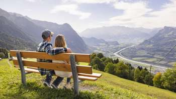 Zwei Wanderer sitzen auf einer Bank und geniessen die Aussicht auf das Rheintal