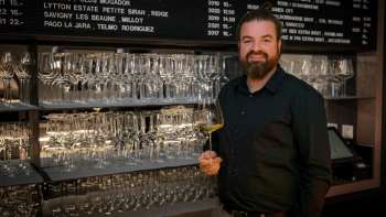 Sympathischer Barkeeper der WY Weinbar mit Weinglas vor stilvollem Regal voller Gläser und Weine.