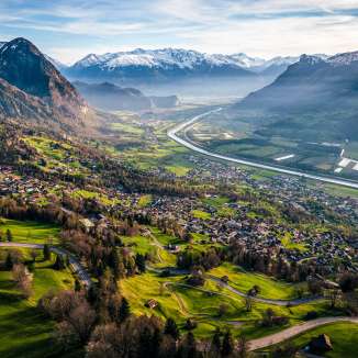 Weitblick vom Hang über das Rheintal mit Feldern, Dörfern und schneebedeckten Bergen – Naturidylle in Liechtenstein