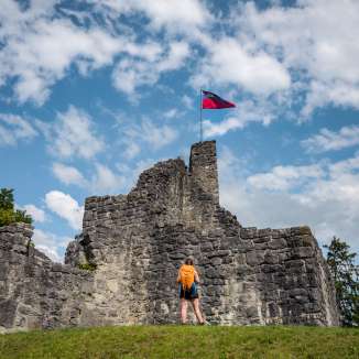 Wanderin vor der historischen Burgruine Schellenberg mit wehender Liechtenstein-Flagge unter blauem Himmel