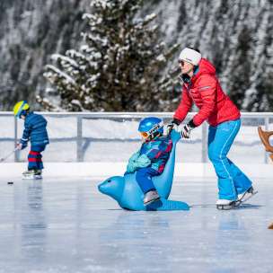 Mutter schiebt Kind auf einem Eislauf-Hilfspinguin über den Eisplatz in Malbun, im Hintergrund ein Kind mit Eishockeyschläger