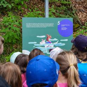 Kindergruppe betrachtet eine Infotafel mit Illustration von „Mila im Wundertunnel“ entlang des Gafadurawegs im Wald.