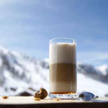 Glas Latte Macchiato auf einer Holzplatte vor der Kulisse verschneiter Berge.