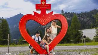 Familie posiert am roten Herzrahmen entlang des Alpherzwegs in Malbun, Liechtenstein, umgeben von alpiner Natur und Bergen.