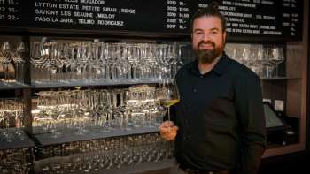 Sympathischer Barkeeper der WY Weinbar mit Weinglas vor stilvollem Regal voller Gläser und Weine.