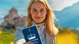 Frau hält den WELCOME Erlebnispass vor Schloss Vaduz in einem blühenden Feld hoch