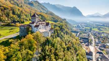 Blick auf die Burg Vaduz mit herbstlich gefärbtem Wald und der Stadt Vaduz im Tal.