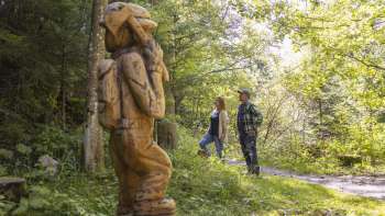 Zwei Wanderer entdecken eine geschnitzte Figur auf dem WalserSagenWeg inmitten eines lichtdurchfluteten Waldes