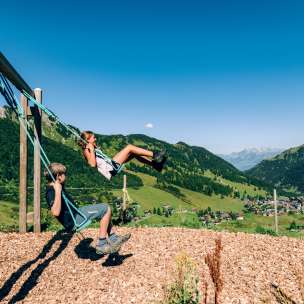 Zwei Kinder schaukeln auf einem Bergspielplatz mit Blick über das Sommerpanorama von Malbun in Liechtenstein