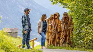 Paar betrachtet geschnitzte Holzfiguren am Walser Sagenweg in Triesenberg mit Blick ins Tal und Bergen im Hintergrund