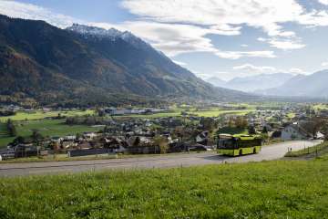 Grüner LIEmobil Bus fährt eine kurvige Straße oberhalb von Liechtenstein entlang mit Blick auf das Rheintal und die umliegenden Alpen