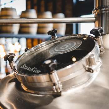 Nahaufnahme eines runden Sichtfensters an einem glänzenden Braukessel mit Blick auf die Sudhalle im Hintergrund.