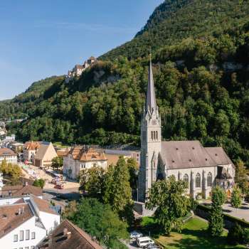 Luftaufnahme der Kathedrale St. Florin in Vaduz mit Schloss Vaduz im Hintergrund an einem sonnigen Tag.