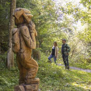 Zwei Wanderer entdecken eine geschnitzte Figur auf dem WalserSagenWeg inmitten eines lichtdurchfluteten Waldes