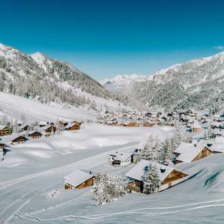 Luftaufnahme von Malbun im Winter mit verschneiten Holzhäusern, eingebettet in die Alpenlandschaft Liechtensteins