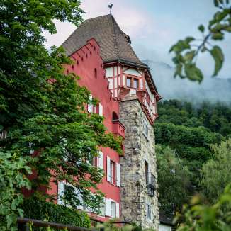 Historisches Rotes Haus in Vaduz, eingebettet in grüne Natur mit Blick auf die umliegenden Berge und einen leicht bewölkten Himmel.