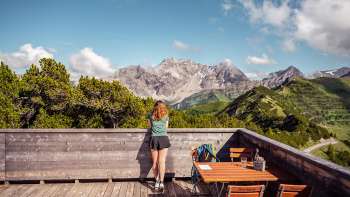 Wanderin blickt von Holzterrasse bei der Aussichtsterasse auf ein beeindruckendes Alpenpanorama.