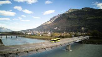 Gelber LIEmobil Bus überquert die Rheinbrücke bei Vaduz mit Bergpanorama im Hintergrund – Liechtenstein erleben
