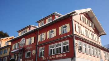 Außenansicht des Alpenhotel Vögeli Malbun mit der Elchbar im Erdgeschoss – rotes Traditionshaus mit weißer Beschriftung vor blauem Himmel. Außenansicht des Alpenhotel Vögeli Malbun mit der Elchbar im Erdgeschoss – rotes Traditionshaus mit weißer Beschriftung vor blauem Himmel.