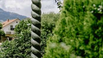 Kunstwerk Renaissance von Daniel Spoerri mit markanter Säulenform und Bronzefigur, umgeben von Natur in Liechtenstein.