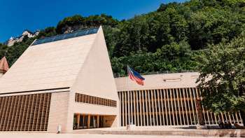 Modernes Landtagsgebäude in Vaduz mit spitzem Giebel, davor die Liechtenstein-Flagge und umgeben von grüner Hügelkulisse.