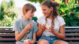 Zwei Kinder sitzen auf einer Parkbank in Liechtenstein und essen Eis – lachende Gesichter, Blumen und Sommerstimmung im Grünen