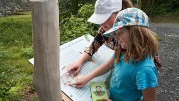 : Kinder stehen vor einer hölzernen Infotafel mit Klapptafeln im Grünen und entdecken spielerisch Wissen entlang des Erlebnispfads „Wurz und Zirp auf Zeitreise“ in Liechtenstein.